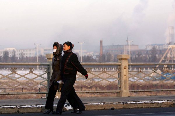 Två kvinnor går på en bro framför fabriker som släpper ut rök.