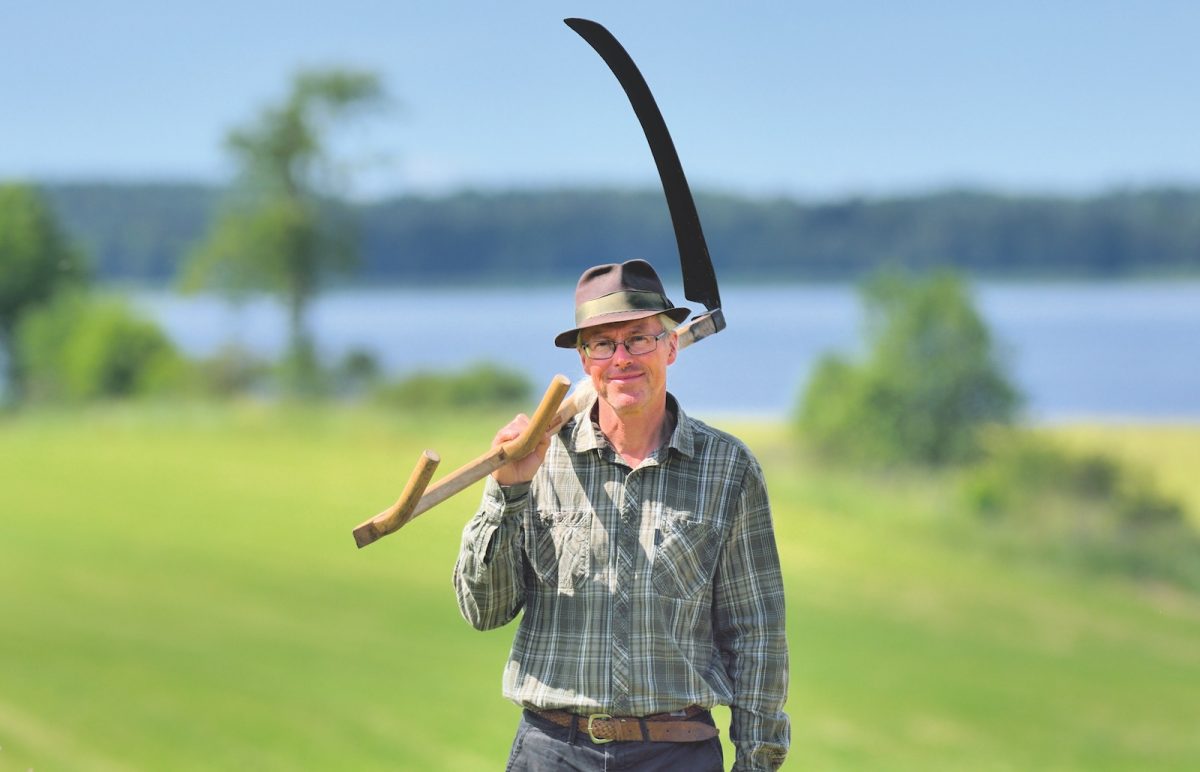 Mats Rosengren i hatt och skjorta med lie framför en nyslagen äng vid Hornborgarsjön.