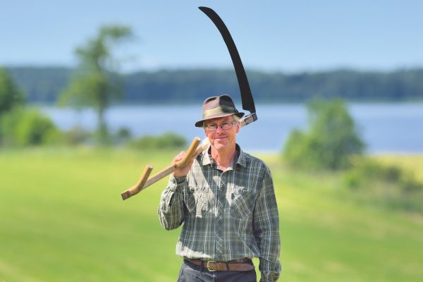 Mats Rosengren i hatt och skjorta med lie framför en nyslagen äng vid Hornborgarsjön.